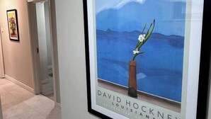 Un pasillo con una obra enmarcada de un paisaje montañoso y un jarrón con flores, con la etiqueta "DAVID HOCKNEY LOUISIANA JANUARY 2002".