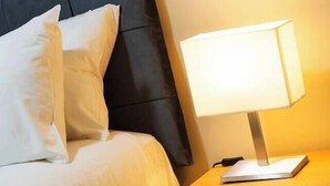 Una cama bien hecha con s ábanas y almohadas blancas, una lámpara de noche y una mesita de noche de madera con cajones.