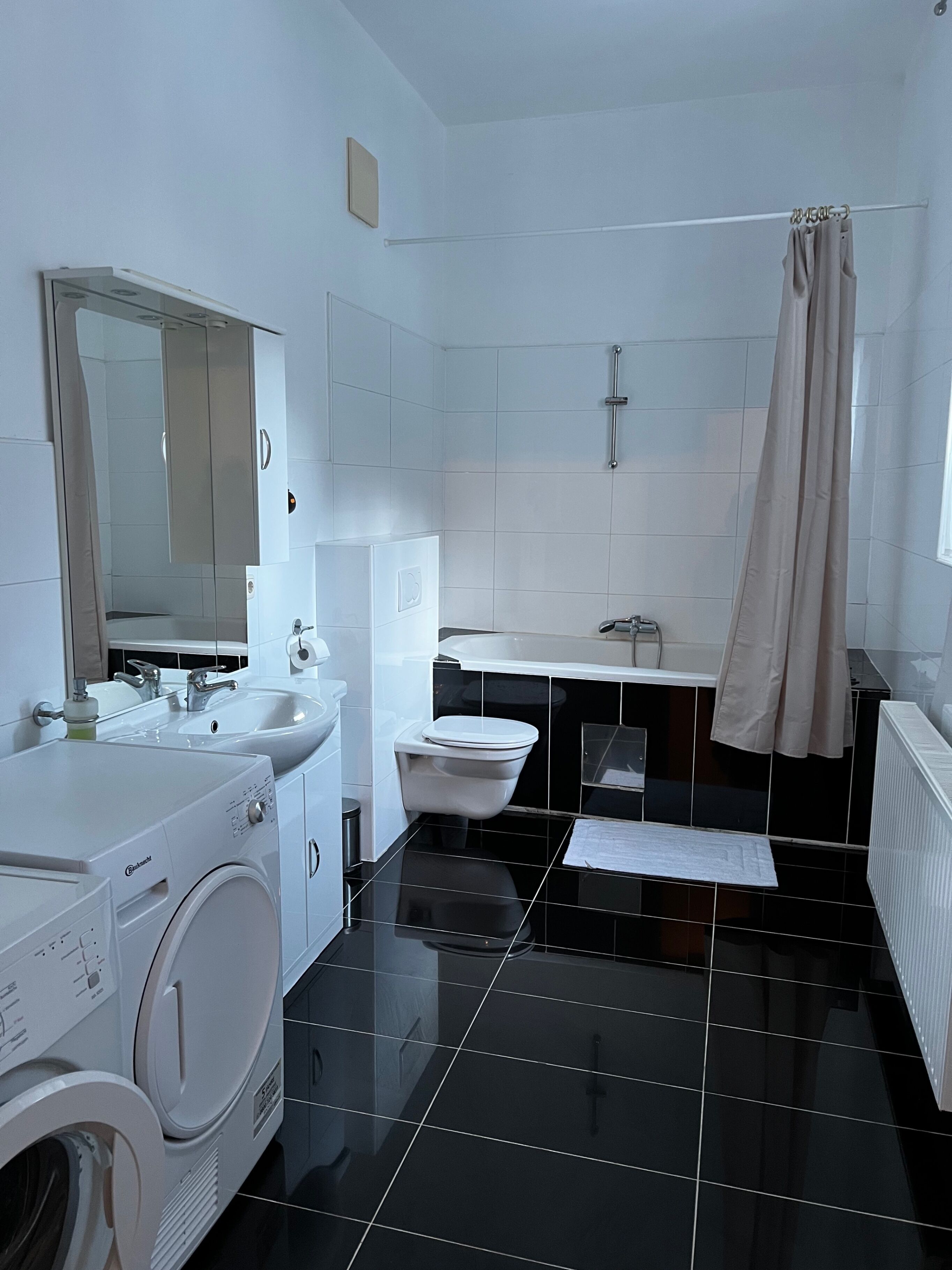 A modern bathroom with a black tiled floor, a white toilet, a sink, a mirror, and a washing machine.