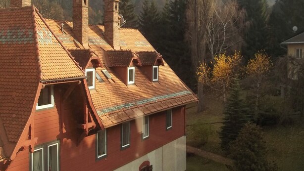 Een huis met twee verdiepingen, met een rode gevel en een pannendak, omgeven door een bos en bergen.
