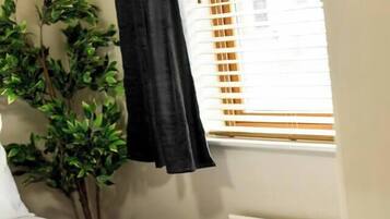 Una cama bien hecha con sábanas blancas y una toalla doblada, una planta en maceta y una ventana con persianas.