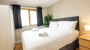Hotellihuone, jossa on suuri sänky, yöpöytä, yövalaisin, ikkuna verhoilla ja ruukkukasvi.