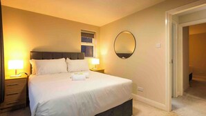 Habitación de hotel con una cama grande, dos mesitas de noche con lámparas, un espejo redondo en la pared y una ventana con persianas.