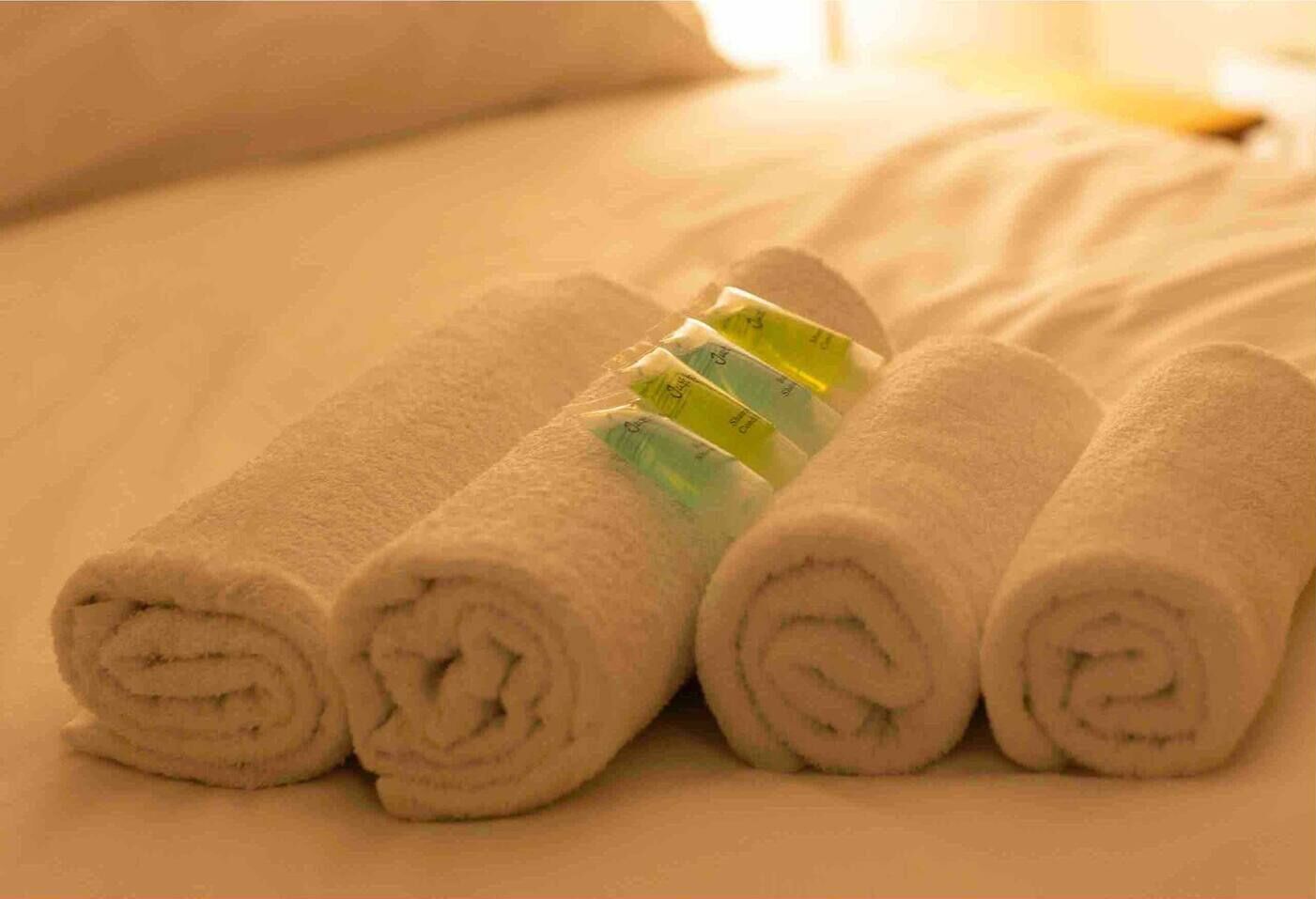 A neatly made bed with rolled white towels and a set of green and yellow toiletries.