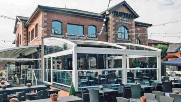 Un espacio al aire libre con mesas y sillas, un patio cubierto y un edificio de ladrillo con un letrero que dice "CUTTERS WHISTLE & BAR".