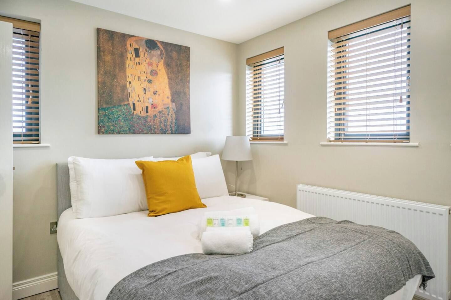 A bedroom with a bed, a painting, and two windows with blinds.