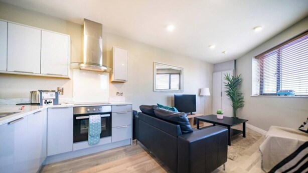 A modern kitchen with white cabinets, a built-in oven, and a stainless steel range hood. There is a black sofa and a small table with a lamp in the living area. - The Manhattan -1bd City Ap (Gold Short Term Stays) (Belfast)