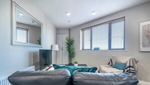 A modern living room with a sofa, a coffee table, a TV, a mirror, and a window with blinds. - The Manhattan -1bd City Ap (Gold Short Term Stays) (Belfast)