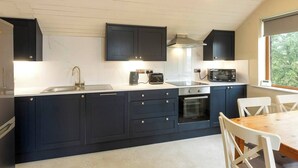 Eine moderne Küche mit dunklen Schränken, einer Spüle aus Edelstahl, einem Toaster, einer Mikrowelle und einem Backofen. Es gibt einen Esstisch mit Stühlen und ein Fenster mit Blick auf Grünflächen.