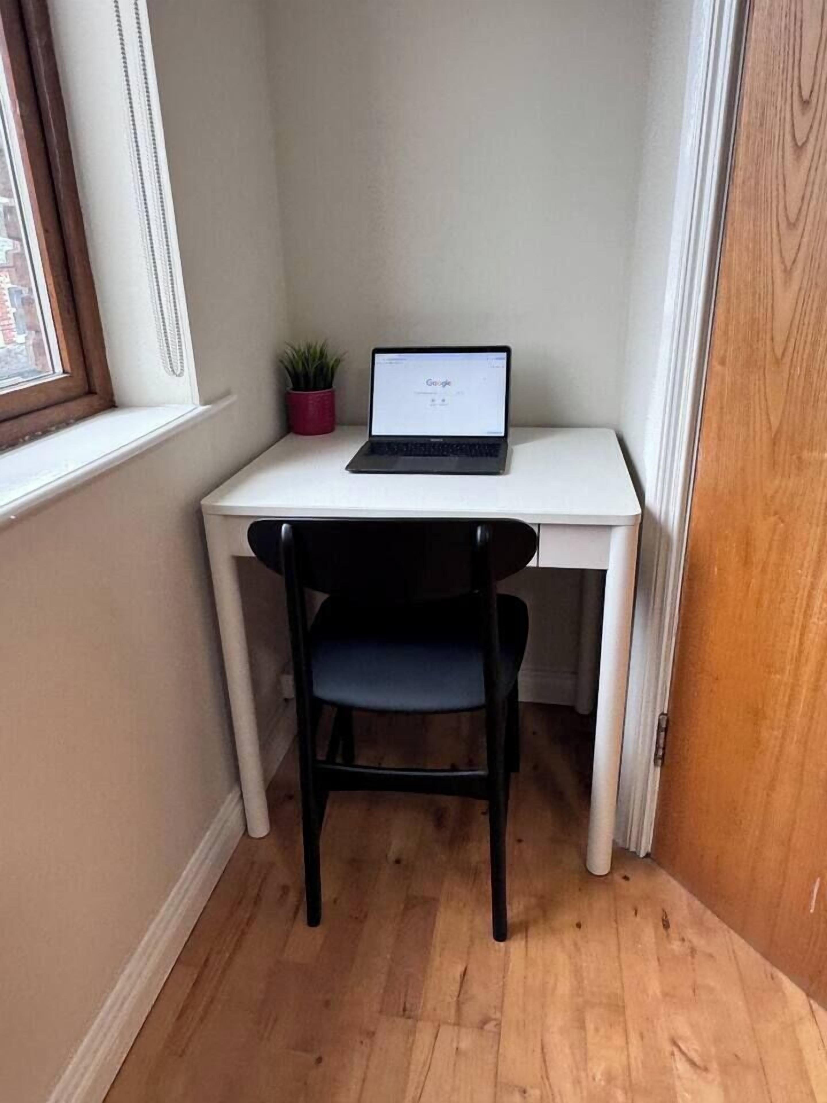 Un pequeño escritorio blanco con una laptop, una silla negra y una planta en maceta en una habitación de esquina.