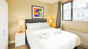 Ein Hotelzimmer mit einem Bett, zwei Nachttischlampen, einem gerahmten abstrakten Kunstwerk, einem Fenster mit Vorhängen und einem Heizkörper.