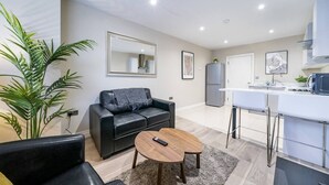 Ein modernes Wohnzimmer mit einer schwarzen Ledersofa, einem Holztisch und einem großen Spiegel. Im Hintergrund ist ein Küchenbereich mit weißen Schränken und einem Kühlschrank zu sehen.