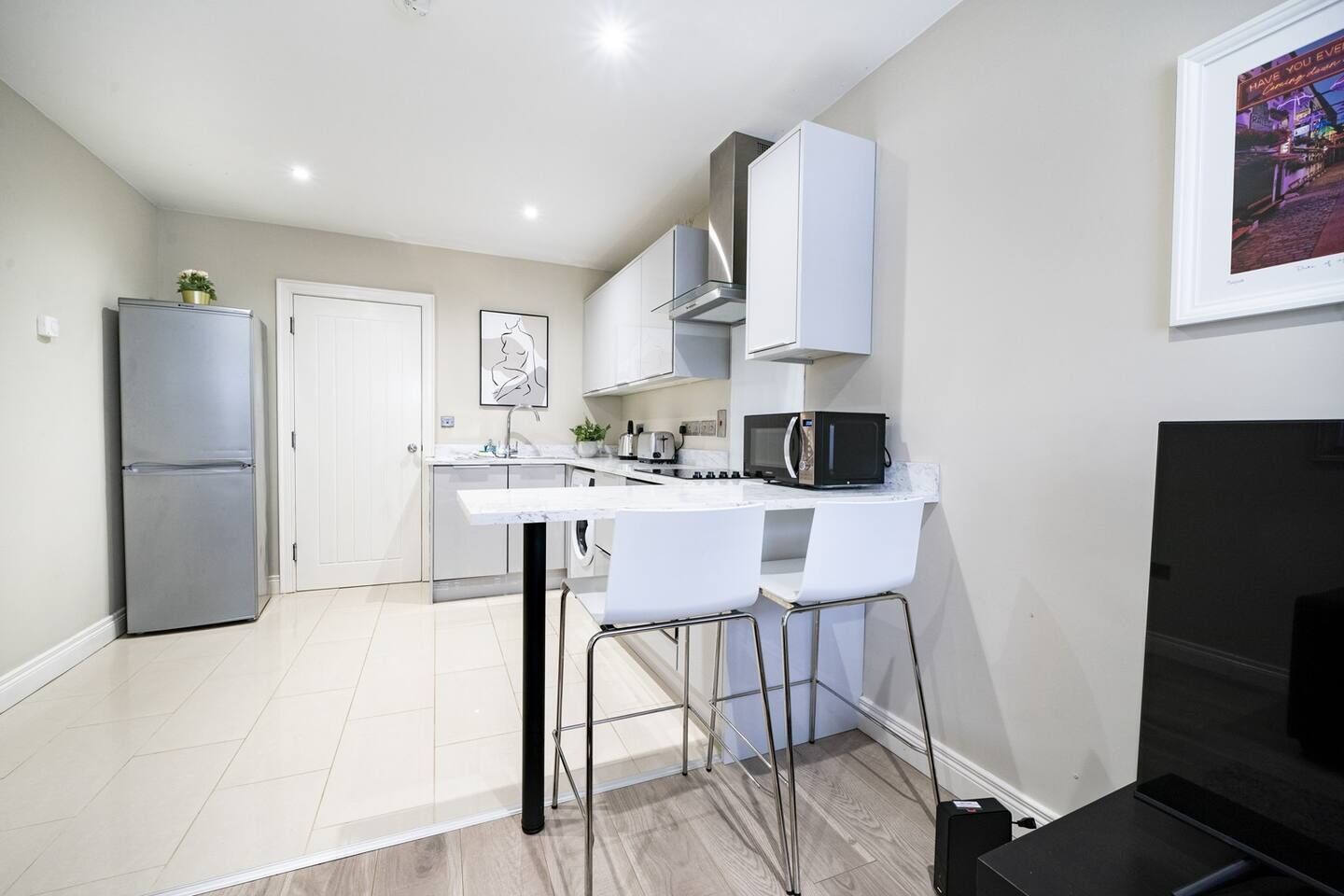 Cocina compacta con encimera blanca, electrodomésticos de acero inoxidable y un taburete de bar.