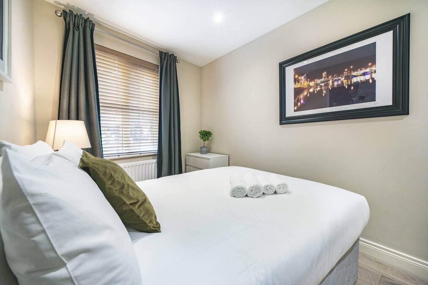 A neatly made bed with white linens and a green pillow, a bedside table with a lamp, a framed picture on the wall, and a window with curtains.