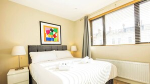Hotelkamer met een bed, twee nachtkastjes, een raam met luxaflex, een radiator en een kleurrijk abstract schilderij aan de muur.