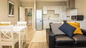 Een compact leefruimte met een kitchenette, een eethoek met witte stoelen en een zwart lederen bank met gestreepte kussens.