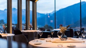 A restaurant with tables set for dining, a vase with flowers, and a view of mountains through large windows.