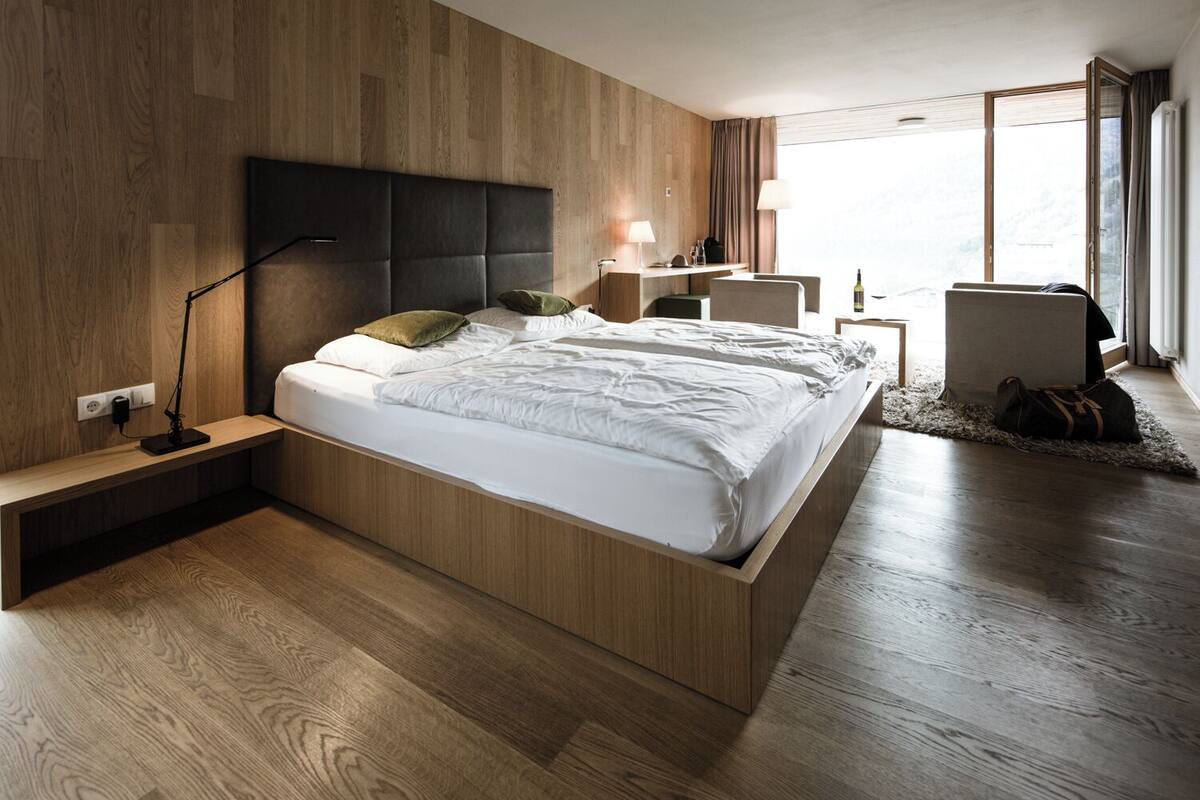 Un dormitorio moderno con cabecera de madera, una mesita de noche con lámpara, un escritorio pequeño y vistas al exterior.