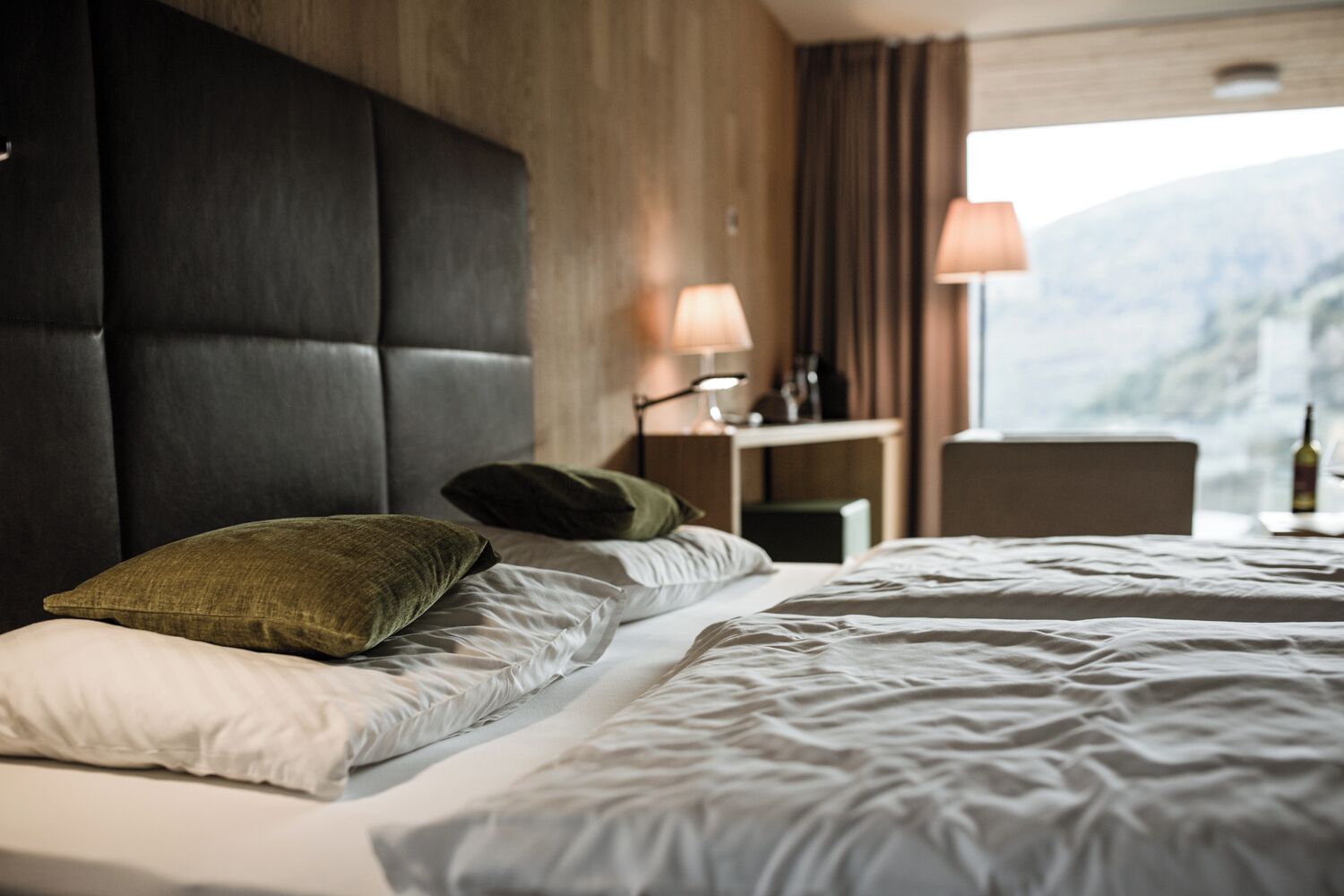 Habitación de hotel con una cama grande, cabecera de madera, mesita de noche con lámpara y vistas a la montaña.