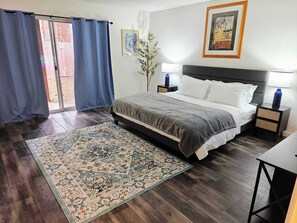 Une chambre à coucher comprenant un lit, deux tables de chevet, une commode, un grand tapis, une fenêtre avec des rideaux et un tableau accroché au mur.