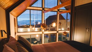 A bedroom with a large window, a bed with pillows, and hanging spherical light fixtures. - Cabanas do Pico / 2A (Canto da Areia)
