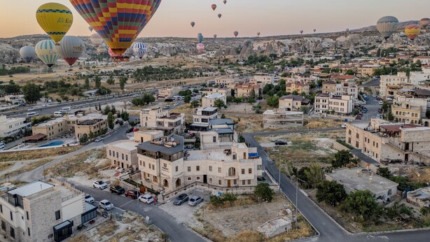 A cityscape with multiple hot air balloons in the sky and a variety of buildings, including a prominent white building with a dome. - Unique Cappadocia Palace (Nevsehir)