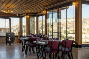 A restaurant with a long table, red chairs, and large windows offering a view of a mountainous landscape. - Unique Cappadocia Palace (Nevsehir)