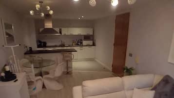 A modern living room with a white sofa, a glass coffee table, and a dining area with a glass table and chairs. A kitchen is visible in the background with white cabinets and a microwave.