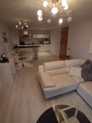 Ein modernes Wohnzimmer mit einem weißen Sofa, einem Glastisch und einem Essbereich mit Tisch und Stühlen. Im Hintergrund ist eine Küche mit weißen Schränken und einer Mikrowelle zu sehen.