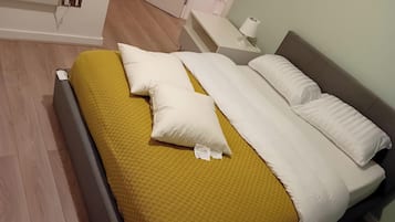A neatly made bed with a yellow blanket and white pillows, a bedside table with a lamp, and a wooden floor.