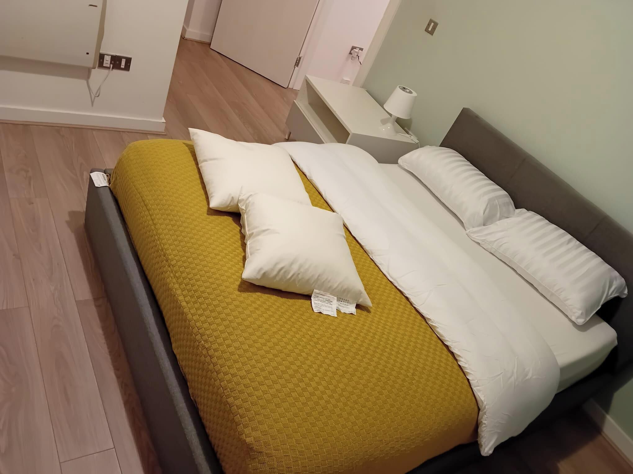 A neatly made bed with a yellow blanket and white pillows, a bedside table with a lamp, and a wooden floor.