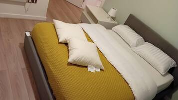 A neatly made bed with a yellow blanket and white pillows, a bedside table with a lamp, and a wooden floor.