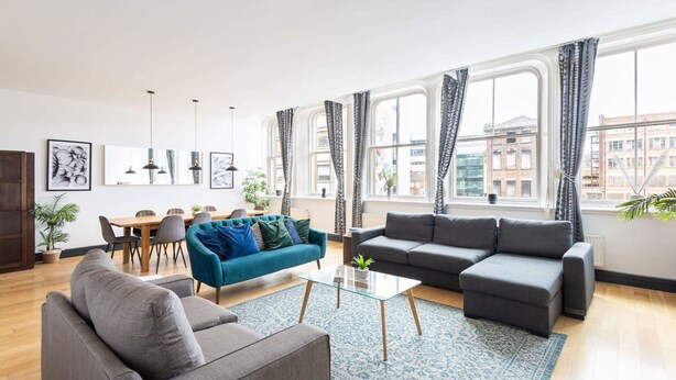 A modern living room with a sofa, a blue armchair, a glass coffee table, and a dining area with chairs and a table. - Host Stay Exchange Street Living (Liverpool)
