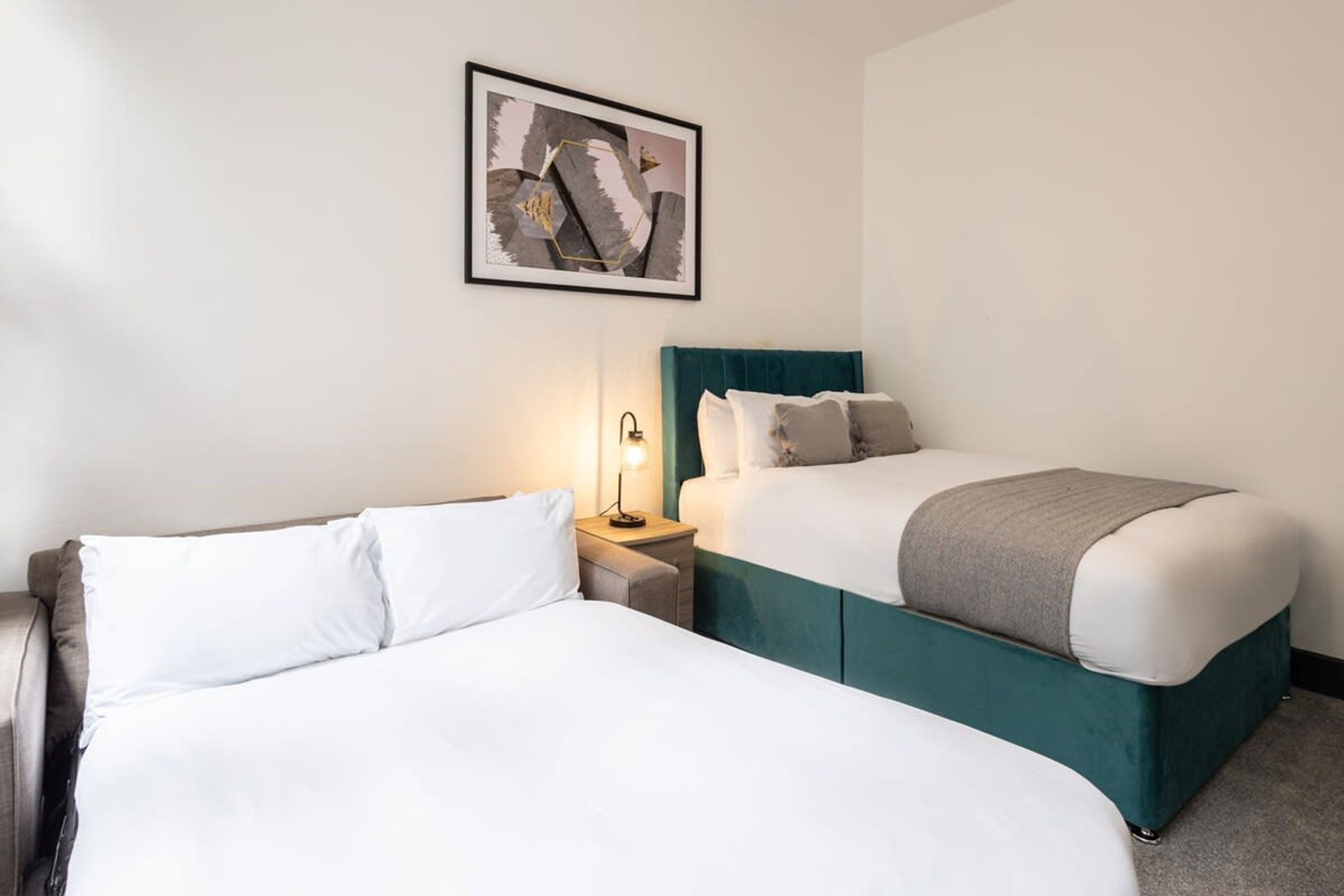 A hotel room with two beds, a nightstand with a lamp, and a framed abstract artwork on the wall.