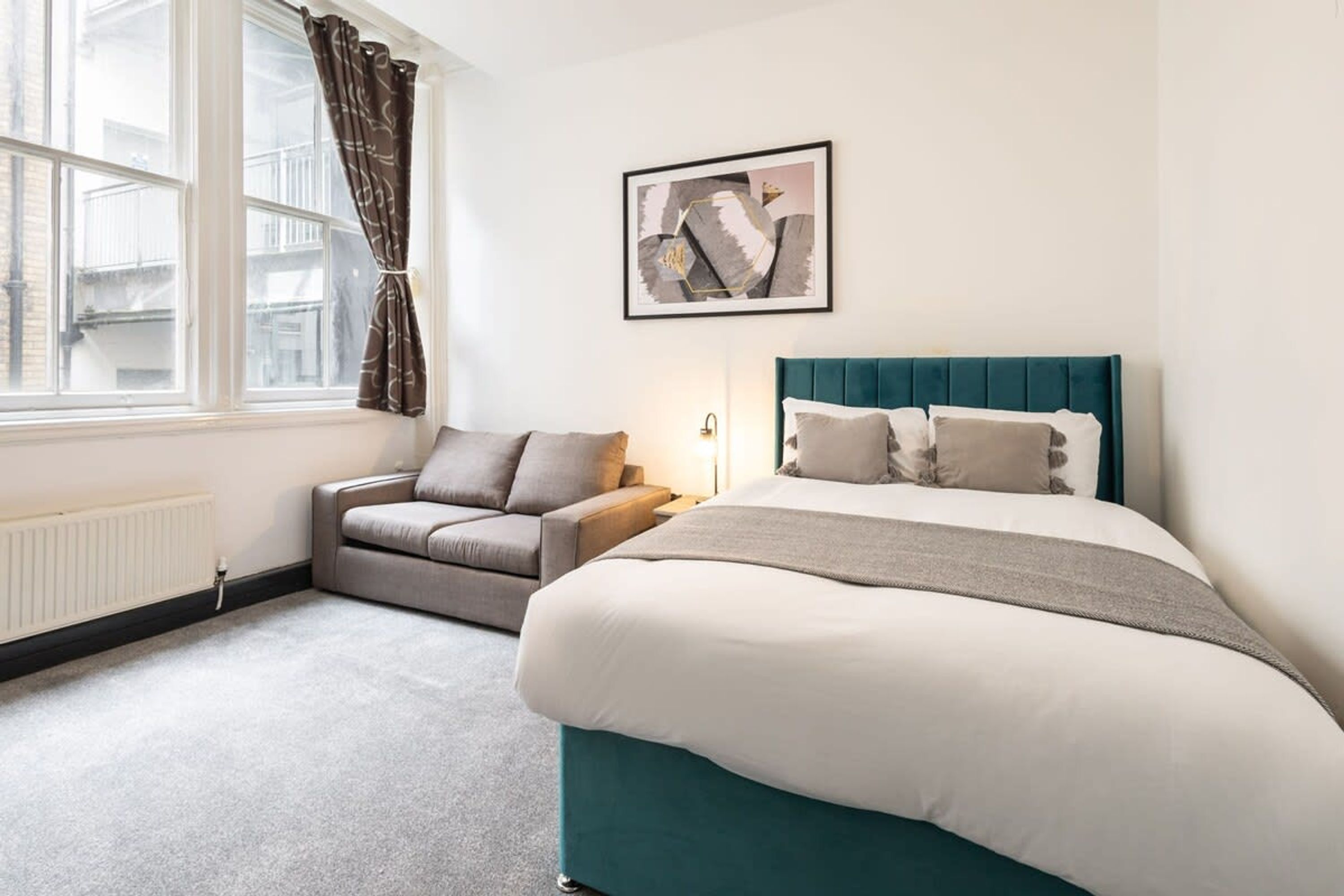 A bedroom with a bed, a sofa, a window with curtains, and a wall-mounted picture.