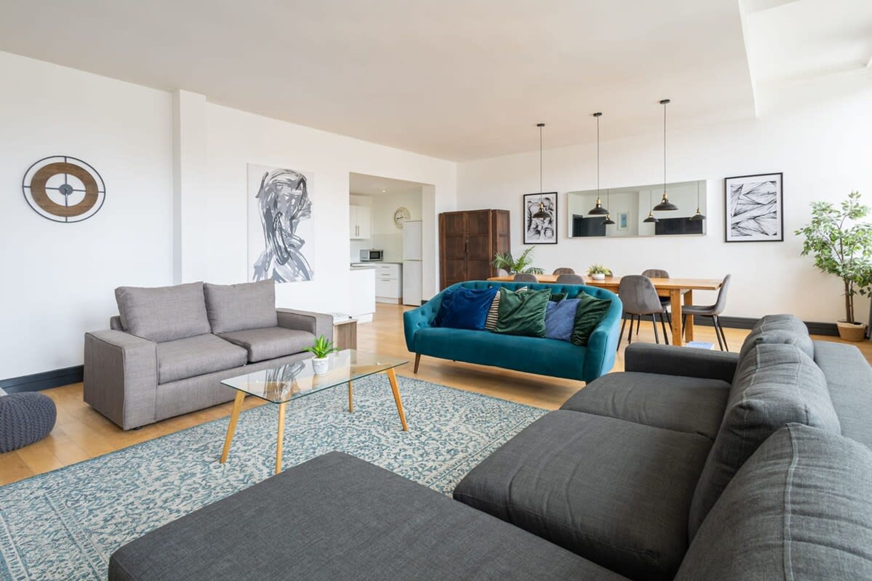En moderne stue med en grå sofa, en tealgrøn sofa og en stor, grå hjørnesofa. Der er et spiseområde med et træbord og stole, samt et køkken i baggrunden.