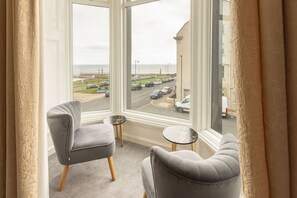 A room with a large window overlooking a parking area and a beach. There are two grey armchairs and a small round table. - Host Stay Glaisdale at the Sandbeck (Whitby)