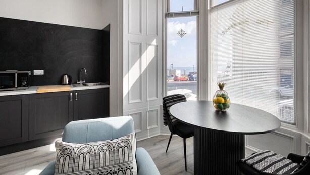 Une cuisine moderne avec des meubles de couleur sombre, un plan de travail noir et un four à micro-ondes. On y trouve un coin repas avec une table ronde et des chaises, ainsi qu’une grande fenêtre équipée de stores.