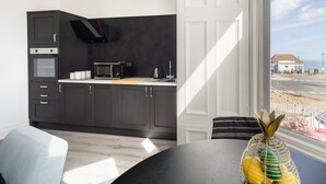 Une cuisine moderne avec des meubles de couleur sombre, un four à micro-ondes et une cuisinière. Une fenêtre offre une vue sur la mer et un immeuble.