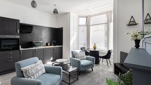 Une cuisine moderne avec des meubles de couleur sombre, un four à micro-ondes et une cuisinière. On y trouve deux fauteuils bleu clair avec des coussins à motifs, une petite table ronde et une table basse. Une grande fenêtre laisse entrer la lumière naturelle, et une plante est visible dans un coin.