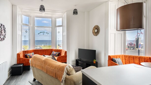 A modern living room with two orange sofas, a beige sofa, a TV, and a large window with a view of the sea.