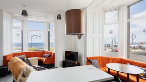 A modern living room with orange sofas, a white table, and a large window with a view of the sea.