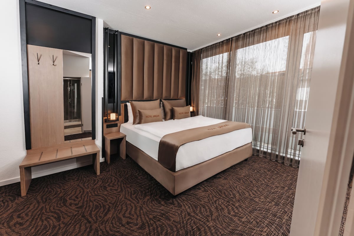 Une chambre d’hôtel moderne avec un grand lit, un banc en bois, un miroir et une grande fenêtre avec des rideaux.