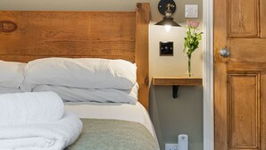 A neatly made bed with white linens and a folded white towel, a bedside table with a lamp and a vase of flowers, and a wooden door with a lock.