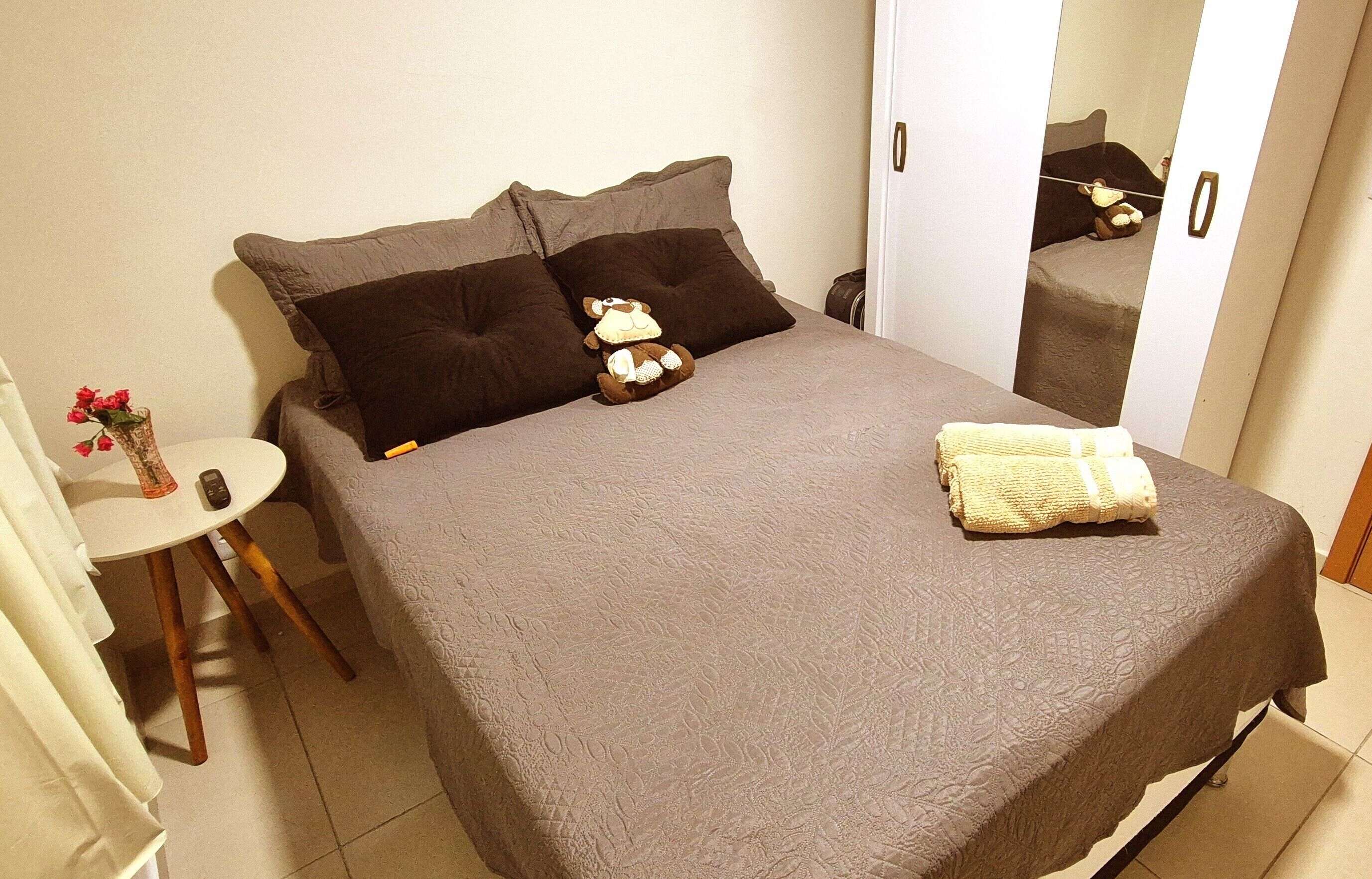A neatly made bed with a grey bedspread, two dark brown pillows, and a small stuffed animal. A white towel is folded on the bed.