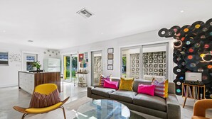 A modern living room with a sofa, a glass coffee table, and a chair. There is a kitchen area in the background. - Palm Springer - Heated Salt Water Pool Steps from the Drive (Wilton Manors)