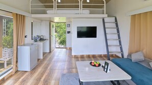 A compact hotel room with a bunk bed, a TV mounted on the wall, a small kitchenette, and a balcony with a view of greenery.