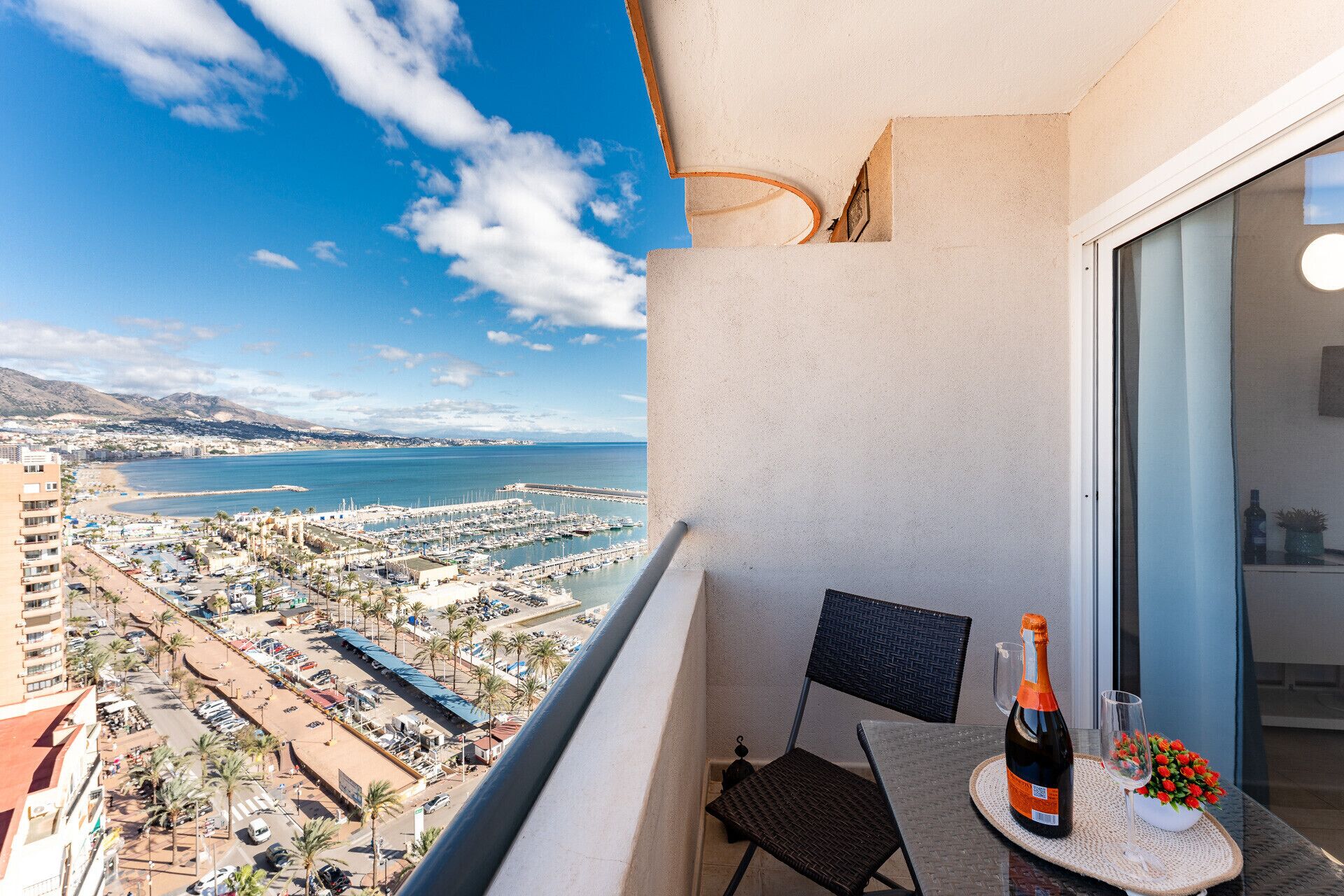 A balcony with a view of a marina and beach, a bottle of champagne, and a chair.