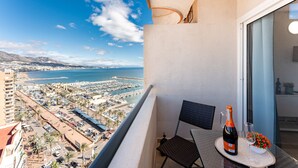 Un balcón con vista a un puerto deportivo y una playa, una botella de champán y una silla.