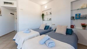 Una habitación limpia y moderna con dos camas, cada una con una toalla azul y una almohada blanca. Hay estantes con artículos de aseo y una planta pequeña.